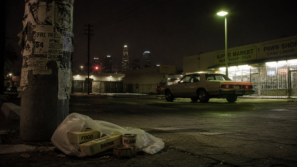 Nighttime 1980s Los Angeles street with anonymous supermarket packaging, punk flyers and a mysterious parked American sedan