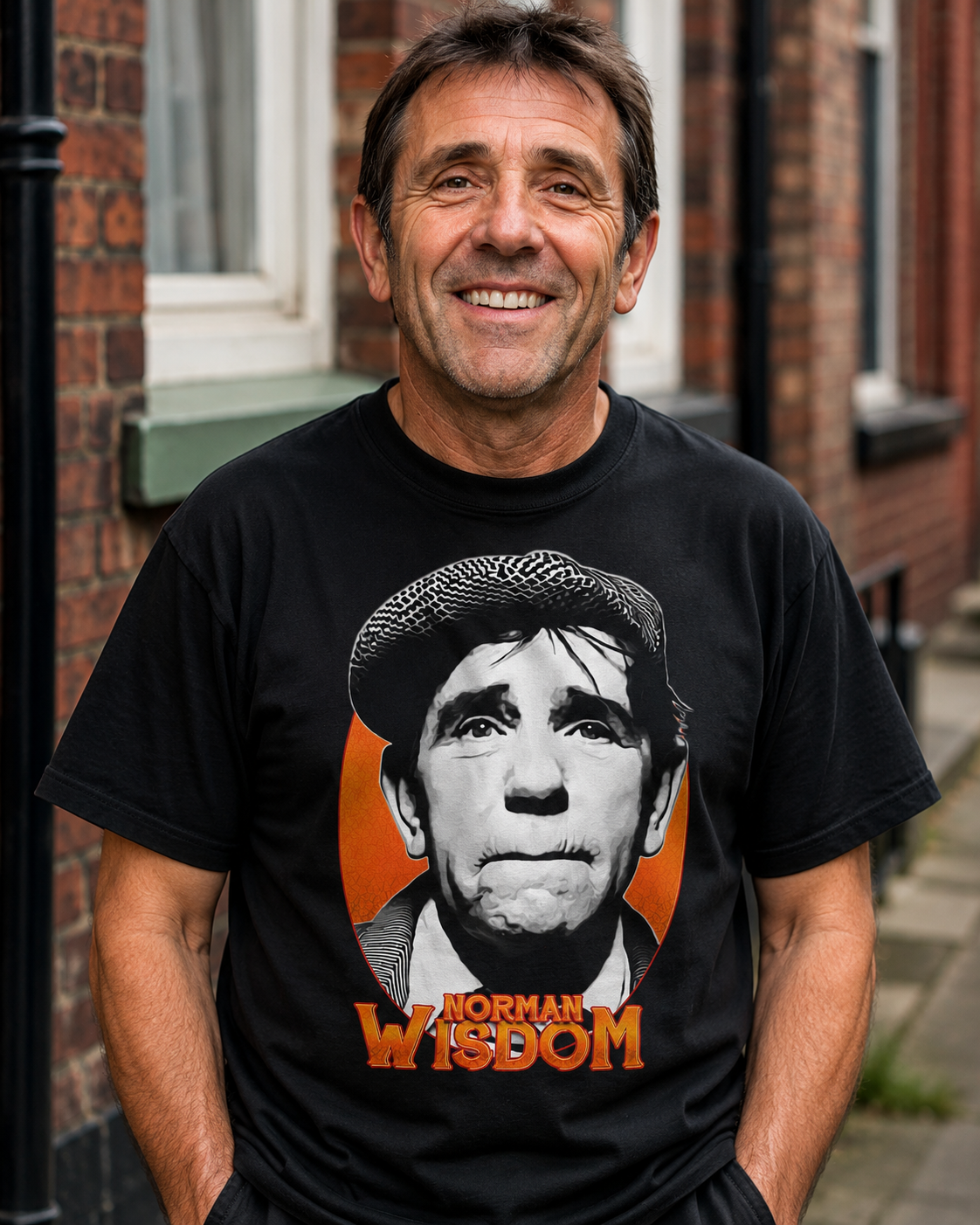 A smiling man wearing a black graphic t-shirt featuring a black and white portrait of Norman Wisdom and the text 'NORMAN WISDOM' in orange, standing outdoors in front of a brick building.